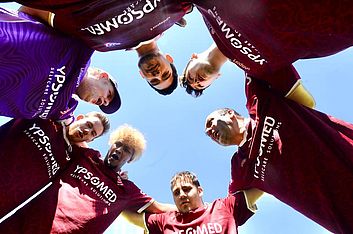 Eine Gruppe von Fußballspielern in kastanienbraunen und violetten Trikots steht in einem Kreis, die Arme umeinander gelegt, und blickt vor dem Hintergrund eines strahlend blauen Himmels in die Kamera.