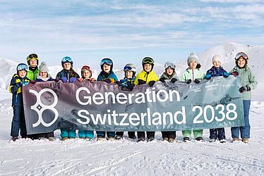 Dreizehn Menschen in Winterkleidung stehen auf verschneitem Boden und halten ein großes Transparent mit der Aufschrift „Generation Schweiz 2038“ hoch, im Hintergrund sind Berge und ein blauer Himmel zu sehen.