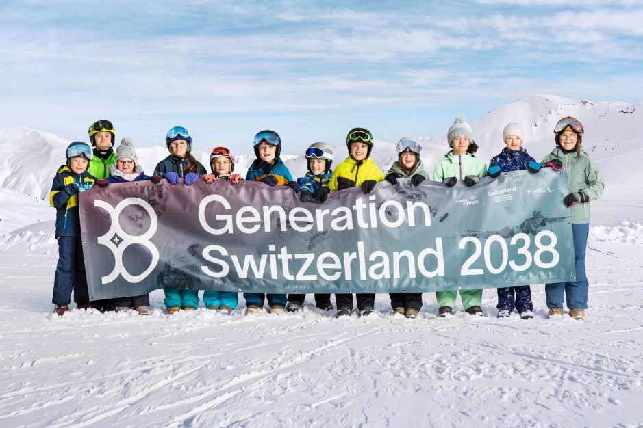 Dreizehn Menschen in Winterkleidung stehen auf verschneitem Boden und halten ein großes Transparent mit der Aufschrift „Generation Schweiz 2038“ hoch, im Hintergrund sind Berge und ein blauer Himmel zu sehen.