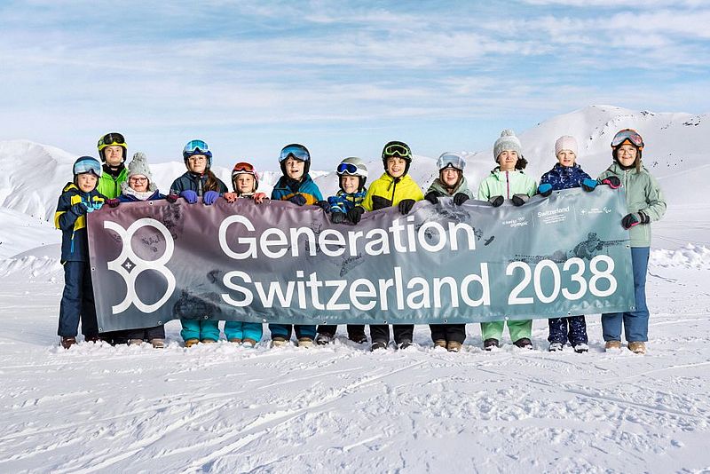 Dreizehn Menschen in Winterkleidung stehen auf verschneitem Boden und halten ein großes Transparent mit der Aufschrift „Generation Schweiz 2038“ hoch, im Hintergrund sind Berge und ein blauer Himmel zu sehen.