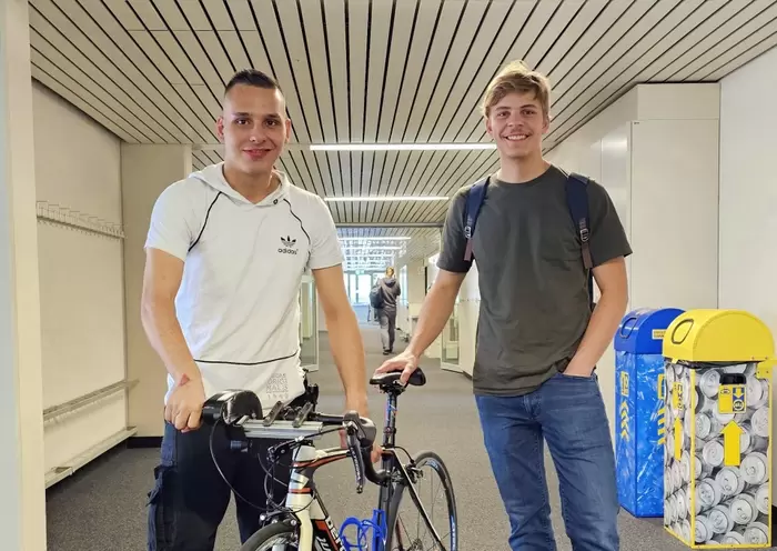 Zwei junge Männer stehen in einem Flur; einer hält ein Fahrrad, beide lächeln in die Kamera. Im Hintergrund sind Recyclingbehälter zu sehen. Der Flur hat weiße Wände und eine gestreifte Decke.