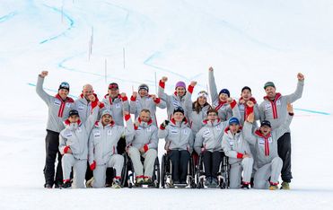Eine Gruppe von Sportlern und Trainern, von denen einige im Rollstuhl sitzen, posieren gemeinsam auf einer verschneiten Piste, tragen passende graue Jacken und Mützen und feiern mit erhobenen Armen und einem Lächeln.