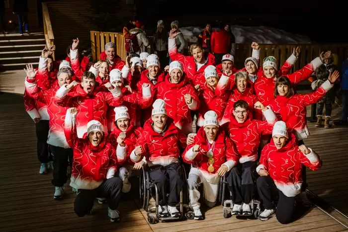 Eine Gruppe von Sportlern in passenden rot-weißen Jacken und Mützen jubelt und hebt die Arme, einige halten Medaillen in der Hand. Zwei Personen in der vorderen Reihe sitzen in Rollstühlen. Die Gruppe steht nachts im Freien auf einer Holzplattform.