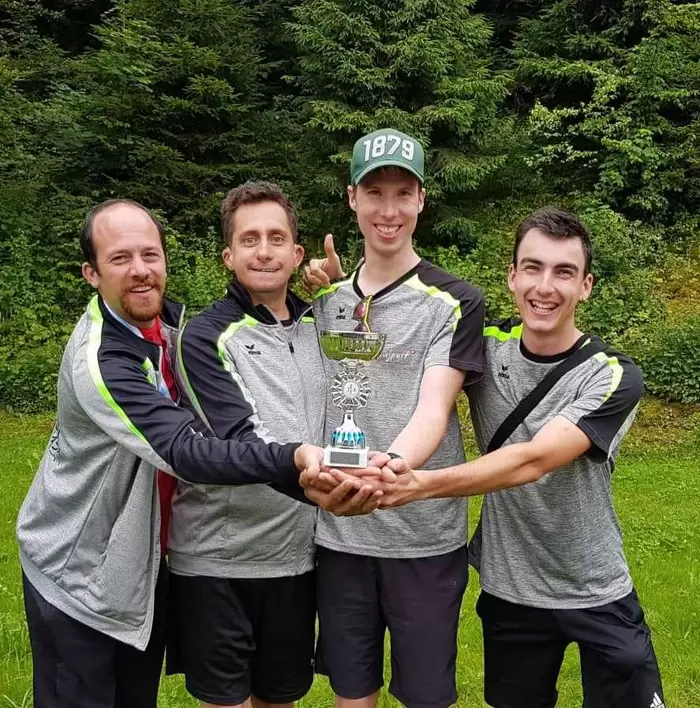 Vier lächelnde Männer in einheitlichen Sporttrikots stehen im Freien und halten gemeinsam einen Pokal in die Höhe. Sie sind von Gras und Bäumen umgeben, und der Mann, der von rechts der Zweite ist, trägt eine grüne Mütze mit der Aufschrift „1879“.