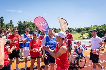 Eine Gruppe von Menschen, darunter eine Person im Rollstuhl, lächelt und feiert auf einer sonnigen Laufbahn im Freien. Einige tragen rote Trikots, und im Hintergrund wehen bunte Transparente. Ein Fotograf hält den Moment fest.