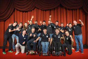 Eine Gruppe von Menschen, einige in Rollstühlen, posiert enthusiastisch vor roten Vorhängen und trägt dazu passende schwarze Hemden mit Logos. Sie lächeln, jubeln und heben feierlich die Arme.