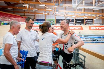 Ein lächelnder Radrennfahrer in Rennkleidung wird von einer Gruppe von Personen in weißen Hemden in einer Radrennhalle beglückwünscht, wobei die Strecke und die leeren Tribünen im Hintergrund zu sehen sind.