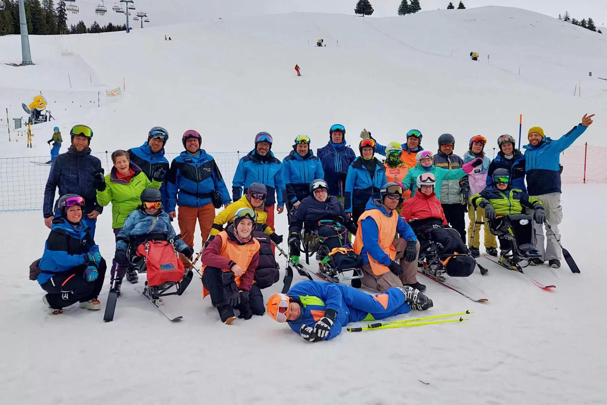 Eine Gruppe von Menschen, einige mit adaptiver Skiausrüstung, posiert zusammen auf einer verschneiten Skipiste. Die meisten tragen Helme und Winterausrüstung und lächeln für das Foto, während im Hintergrund Skilifte und Pisten zu sehen sind.