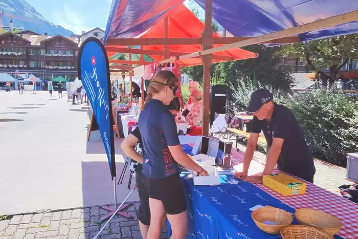 Eine Person steht an einem Marktstand im Freien und unterhält sich unter einem roten Vordach mit einem Verkäufer. In der Nähe stehen Körbe, eine Kasse und bunte Marktstände. Im Hintergrund sind Gebäude und Berge zu sehen.