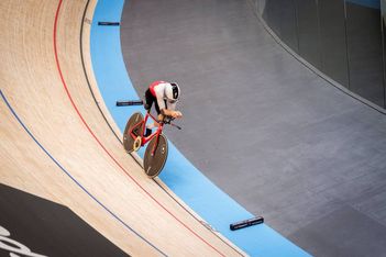 Ein Radfahrer in einem weiß-roten Anzug fährt mit einem Rennrad auf einer Indoor-Radrennbahn, wobei er sich in einer aerodynamischen Position nahe der gekrümmten Kante der Bahn nach vorne lehnt.