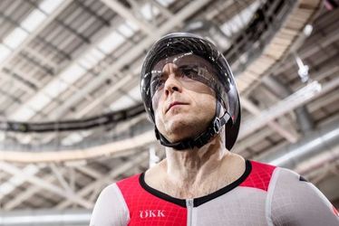 Ein männlicher Bahnradfahrer in rot-weißer Schweizer Uniform und Helm steht unter dem hellen Dach einer Velodromhalle und sieht konzentriert und rennbereit aus.
