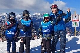 Vier Personen in Skikleidung posieren auf einem schneebedeckten Berg, im Hintergrund sind der blaue Himmel und Berge zu sehen. Drei von ihnen tragen blaue „Ara Ski Training“-Westen. Rechts ist ein Schild mit der Abbildung eines Skifahrers und der Aufschrift „STOP“ zu sehen.