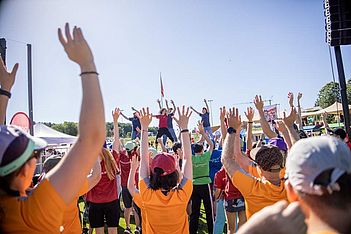 Bei einer Veranstaltung an einem sonnigen Tag hebt eine Gruppe von Menschen im Freien die Hände und folgt dabei den Anweisungen der Trainer auf der Bühne. Die meisten tragen bunte T-Shirts und Hüte. Im Hintergrund sind Zelte und Bäume zu sehen.