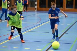 Zwei Jungen spielen Hallenfußball auf einem blauen Platz. Ein Junge in einer blauen Uniform dribbelt einen gelben Ball, während ein Junge in einer grünen Weste versucht, ihn zu verteidigen. Im Hintergrund sind andere Kinder und ein Tor zu sehen.