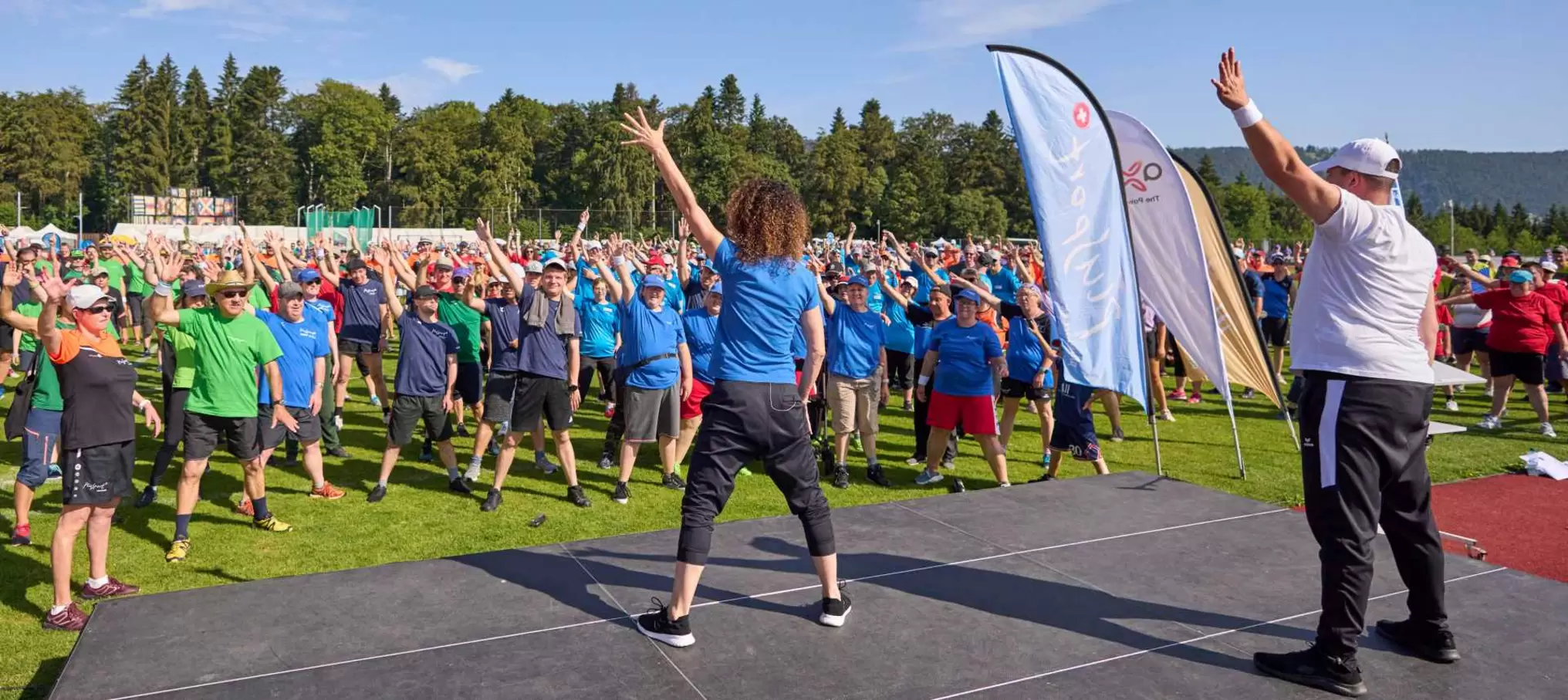 Eine große Gruppe von Menschen in farbenfrohen Hemden nimmt an einer Gruppenübung im Freien teil. Sie folgen zwei Trainern auf einer Bühne, mit Bäumen und strahlend blauem Himmel im Hintergrund.
