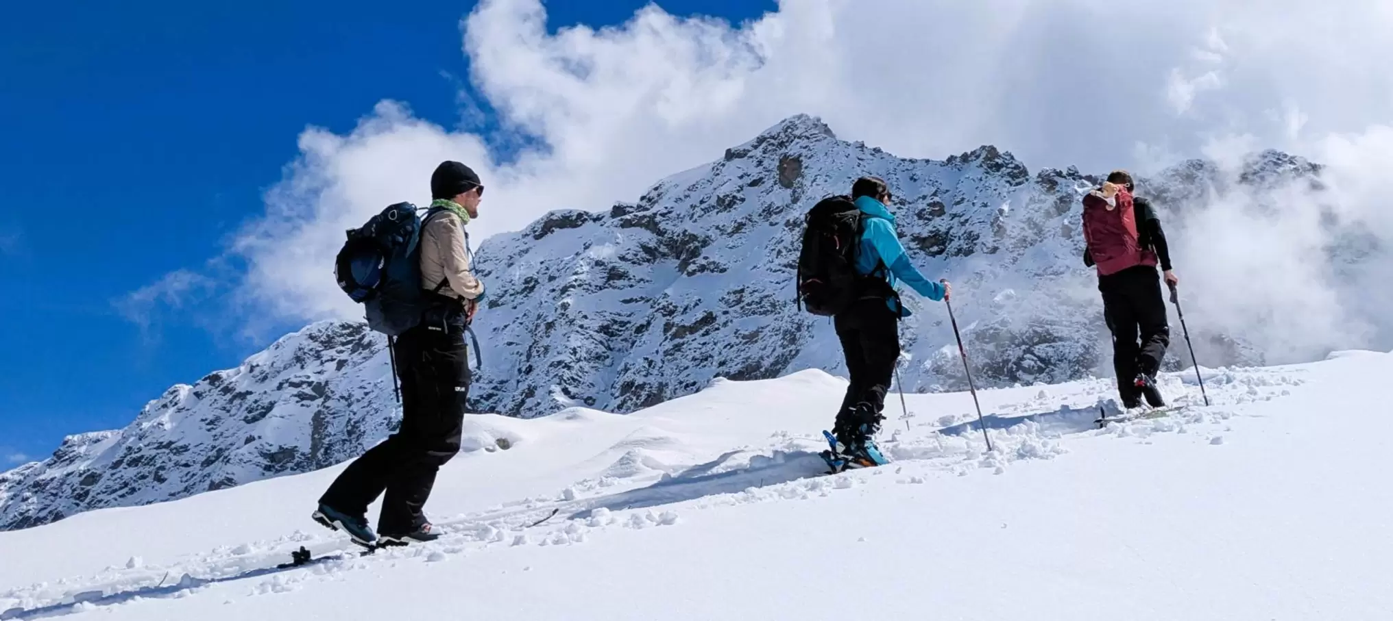 Drei Personen in Winterausrüstung und mit Rucksäcken wandern mit Trekkingstöcken einen verschneiten Berghang hinauf, mit schneebedeckten Gipfeln und Wolken im Hintergrund.