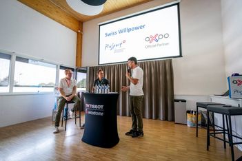 Drei Personen befinden sich in einem hell erleuchteten Raum, zwei sitzend und eine stehend und sprechend, neben einem Tisch mit Wasserflaschen. Ein Bildschirm hinter ihnen zeigt die Logos von Swiss Willpower, PluSport und Axpo.
