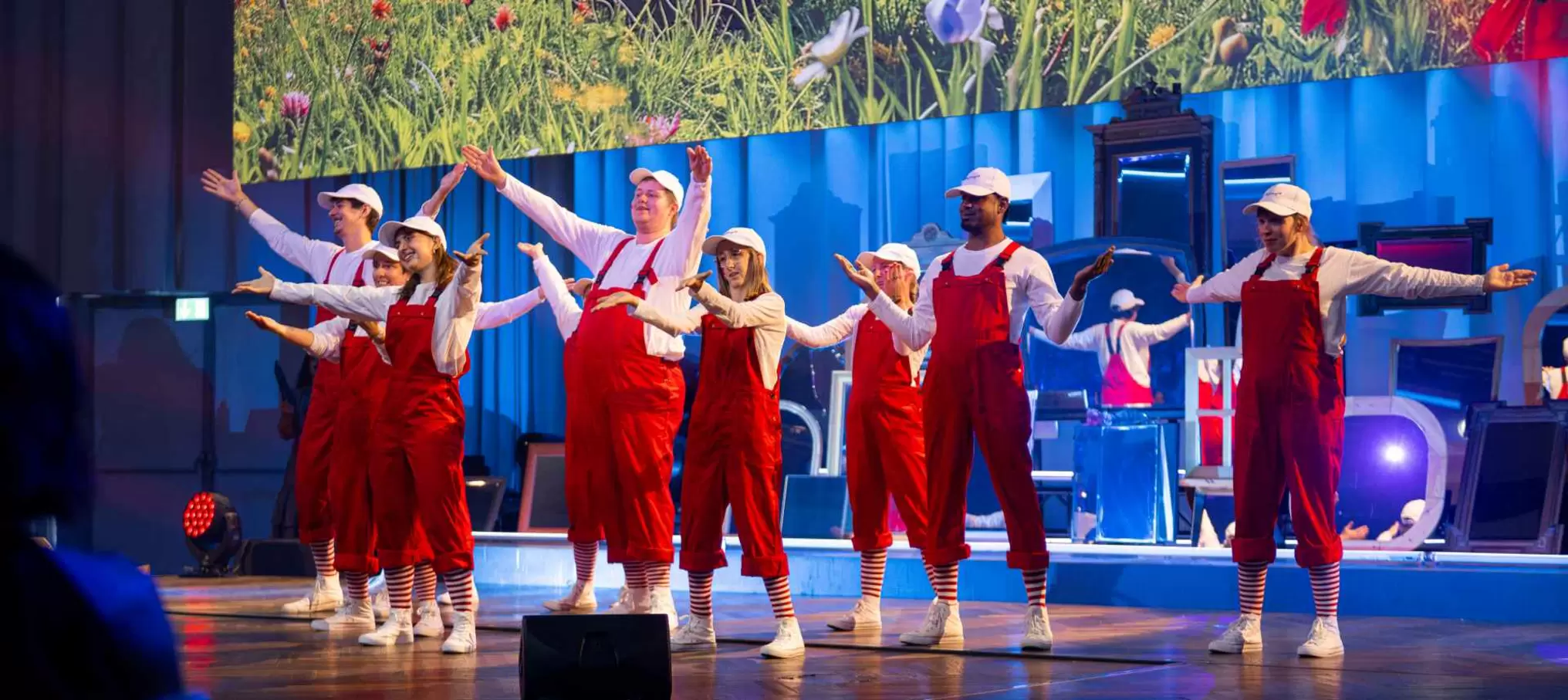 Eine Gruppe von Darstellern in roten Overalls, weißen Hemden, gestreiften Socken und weißen Mützen steht mit ausgestreckten Armen auf der Bühne vor einem bunten Blumenhintergrund und in blauem Licht.