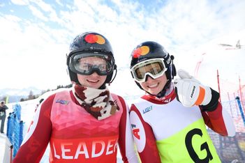 Zwei lächelnde Alpinskifahrer mit Helmen und Skibrillen posieren im Freien auf einer verschneiten Piste. Der Skifahrer auf der linken Seite trägt eine rote LEADER-Weste, während der Skifahrer auf der rechten Seite eine weiß-grüne Weste trägt und einen Daumen nach oben zeigt.