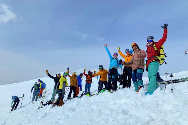 Eine Gruppe von Menschen in farbenfroher Winterkleidung steht mit Skistöcken auf einer verschneiten Piste, lächelt und hebt feierlich die Arme unter einem teilweise bewölkten Himmel.