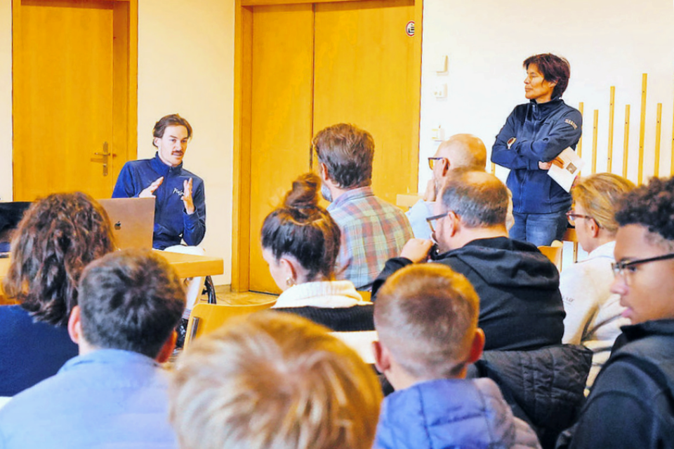 Eine Person sitzt an einem Tisch und spricht zu einer aufmerksamen Gruppe von Menschen in einem Raum. Eine weitere Person steht in der Nähe, hört zu und hält Papiere in der Hand. Die Zuhörer sitzen dem Redner auf Stühlen gegenüber.