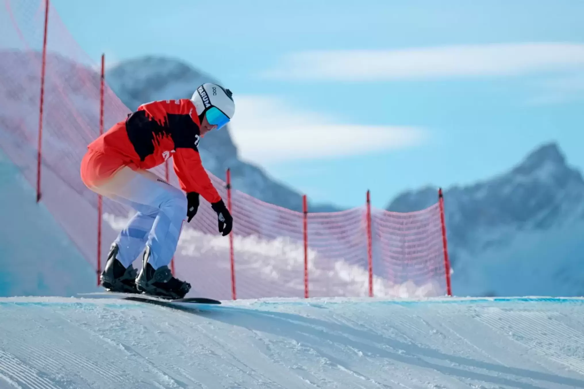 Ein Snowboarder in einem rot-weißen Anzug und einer blauen Brille fährt eine verschneite Piste hinunter, mit einem roten Netzzaun und schneebedeckten Bergen im Hintergrund unter einem klaren Himmel.