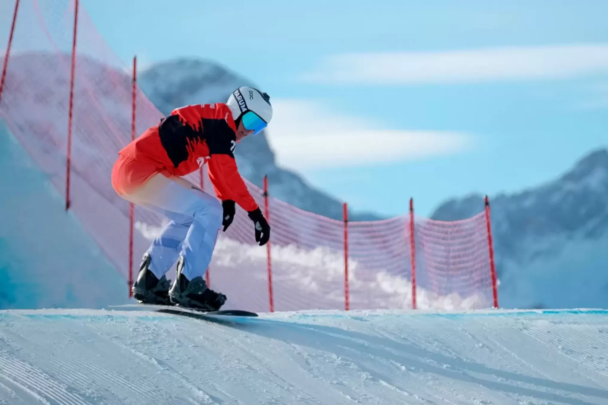 Ein Snowboarder in einem rot-weißen Anzug und einer blauen Brille fährt eine verschneite Piste hinunter, mit einem roten Netzzaun und schneebedeckten Bergen im Hintergrund unter einem klaren Himmel.