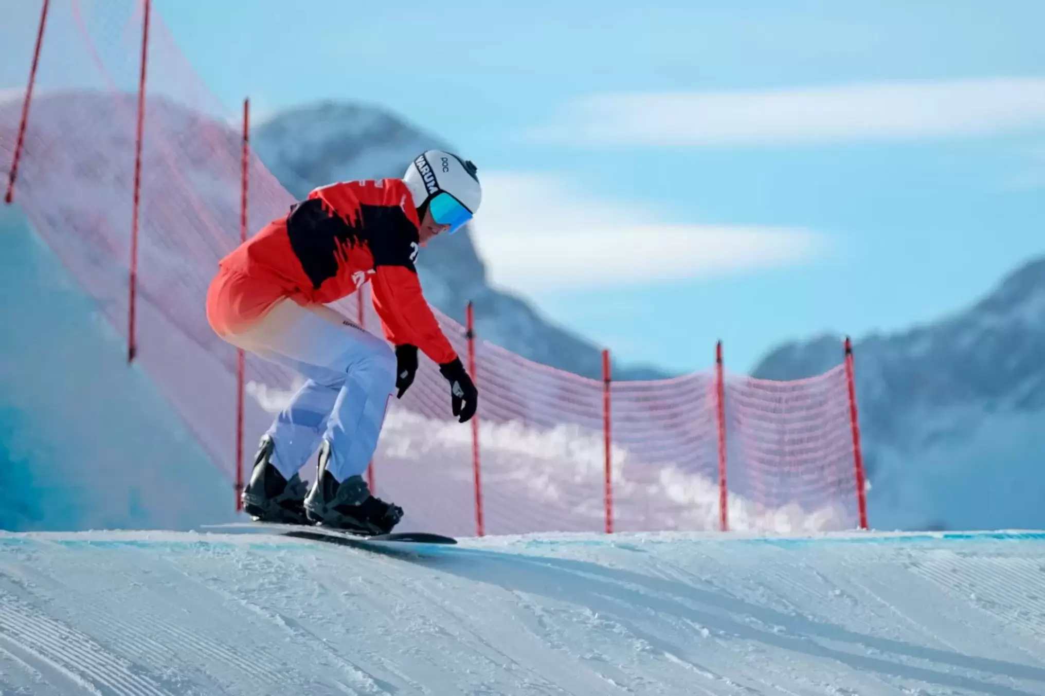Ein Snowboarder in einem rot-weißen Anzug und einer blauen Brille fährt eine verschneite Piste hinunter, mit einem roten Netzzaun und schneebedeckten Bergen im Hintergrund unter einem klaren Himmel.