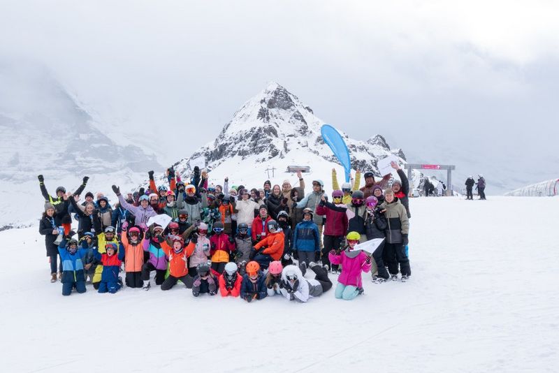 Eine große Gruppe von Menschen, darunter Kinder und Erwachsene in bunter Winterkleidung, posieren zusammen auf einem verschneiten Berg mit Skiausrüstung. Im Hintergrund sind schneebedeckte Gipfel und bewölkter Himmel zu sehen.