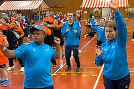 Eine Gruppe von Personen in Sportkleidung nimmt an einer Gruppenaktivität oder einem Aufwärmtraining in einer Sporthalle mit gestreiften Zelten im Hintergrund teil. Alle scheinen konzentriert zu sein, einige heben die Arme und bewegen sich energisch.