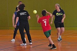 Vier Personen spielen auf einem orangefarbenen Platz ein Hallenspiel. Ein Kind in einem roten T-Shirt ist gerade dabei, einen gelben Ball zu werfen, während drei Erwachsene zusehen und sich darauf vorbereiten, zu reagieren.