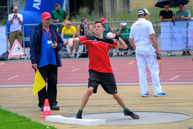 Ein junger männlicher Athlet in einer rot-schwarzen Uniform bereitet sich auf das Kugelstoßen bei einer Leichtathletikveranstaltung im Freien vor, während Offizielle und Zuschauer im Hintergrund zuschauen.
