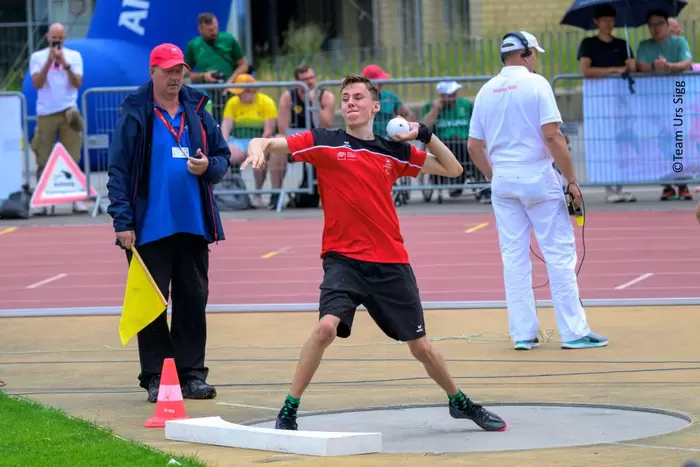 Ein junger männlicher Athlet in einer rot-schwarzen Uniform bereitet sich auf das Kugelstoßen bei einer Leichtathletikveranstaltung im Freien vor, während Offizielle und Zuschauer im Hintergrund zuschauen.