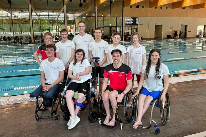 Eine Gruppe junger Schwimmer, einige im Rollstuhl, andere stehend, posiert gemeinsam neben einem Hallenbad. Sie lächeln und tragen Sportkleidung. Im Hintergrund ist eine moderne Schwimmanlage zu sehen.
