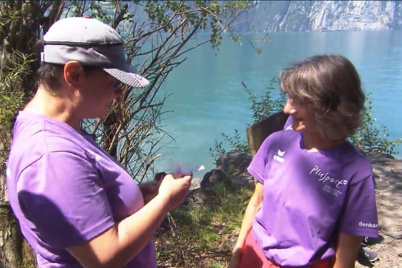 Zwei Frauen in lilafarbenen Hemden stehen an einem Seeufer. Die eine schaut auf etwas in ihren Händen, während die andere sie anlächelt. Im Hintergrund sind Bäume und ruhiges blaues Wasser zu sehen, auf der anderen Seite des Sees sind felsige Klippen zu erkennen.