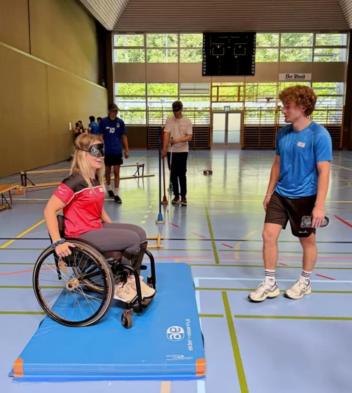 Eine Frau im Rollstuhl, die eine Augenbinde trägt, sitzt auf einer blauen Matte in einer Turnhalle, während ein Mann daneben steht. Im Hintergrund nehmen weitere Personen an Aktivitäten teil.