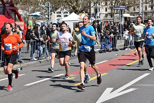 Läuferinnen und Läufer nehmen an einem sonnigen Tag an einem Straßenrennen teil, während Zuschauer hinter Absperrungen zusehen und anfeuern. Einige Läufer tragen nummerierte Startnummern und Sportkleidung, und die Straße ist mit weißen und roten Linien markiert.