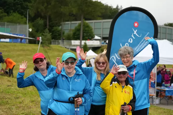 Fünf lächelnde Menschen, vier in blauen Jacken und Hüten und einer in einer gelben Jacke, winken der Kamera in einem Außenbereich mit Gras, Zelten und einem großen PlaySport-Banner hinter ihnen zu.