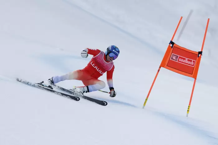Ein Skifahrer in einem roten Anzug und mit blauem Helm macht eine scharfe Kurve um ein orangefarbenes Tor mit der Aufschrift Saalbach auf einer verschneiten Abfahrtsstrecke.