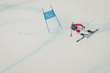 Ein Skifahrer in rot-weißem Anzug und Helm fährt während eines Wettkampfs auf einer verschneiten Piste an einer blauen Fahne mit dem paralympischen Symbol vorbei bergab.