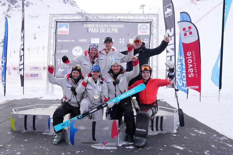 Eine Gruppe von Para-Alpin-Skisportlern und Trainern, einige sitzend, andere stehend, posieren und feiern gemeinsam auf einer verschneiten Piste vor einem "Para Ski Alpin"-Veranstaltungsbanner, wobei Skiausrüstung und Nationalflaggen sichtbar sind.