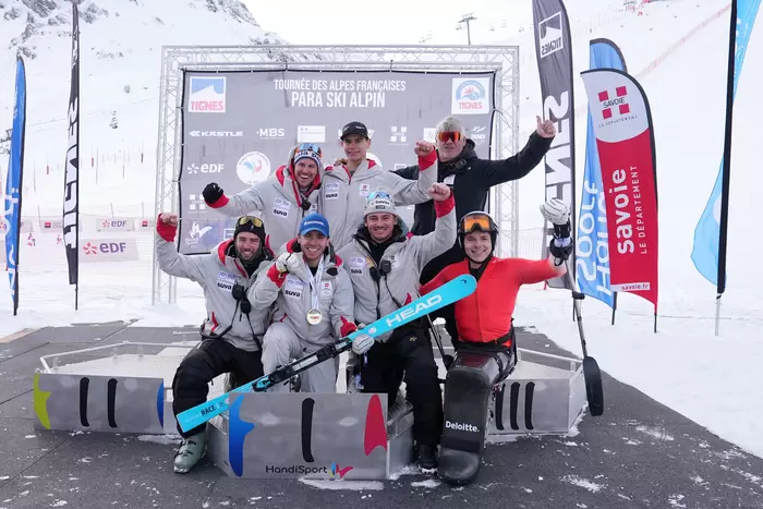 Eine Gruppe von Para-Alpin-Skisportlern und Trainern, einige sitzend, andere stehend, posieren und feiern gemeinsam auf einer verschneiten Piste vor einem "Para Ski Alpin"-Veranstaltungsbanner, wobei Skiausrüstung und Nationalflaggen sichtbar sind.