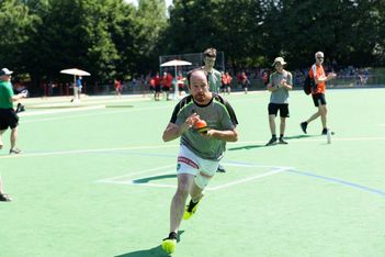 Ein Mann in Sportkleidung rennt energisch auf einem grünen Sportplatz, in der Hand eine orangefarbene Schultüte. Mehrere Menschen schauen zu oder laufen im Hintergrund unter sonnigem Himmel, umgeben von Bäumen.