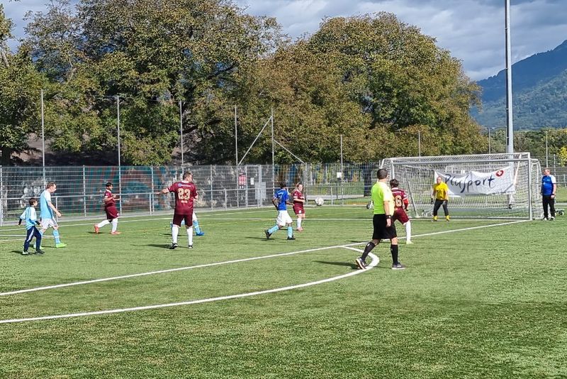 Ein Fußballspiel findet auf einem von Bäumen und Bergen umgebenen Feld statt. Die Spieler in kastanienbraunen und blauen Trikots befinden sich in der Nähe des Tores, während ein Schiedsrichter in Gelb das Spiel aufmerksam beobachtet. Der Himmel ist teilweise bewölkt.