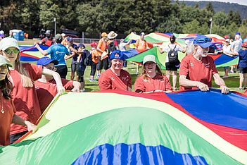 Eine Gruppe von Menschen in bunten Hemden und Hüten hält an einem sonnigen Tag im Freien einen großen, bunten Fallschirm hoch und nimmt an einer Gruppenaktivität auf einer Wiese teil. Im Hintergrund sind Bäume und andere Gruppen zu sehen.