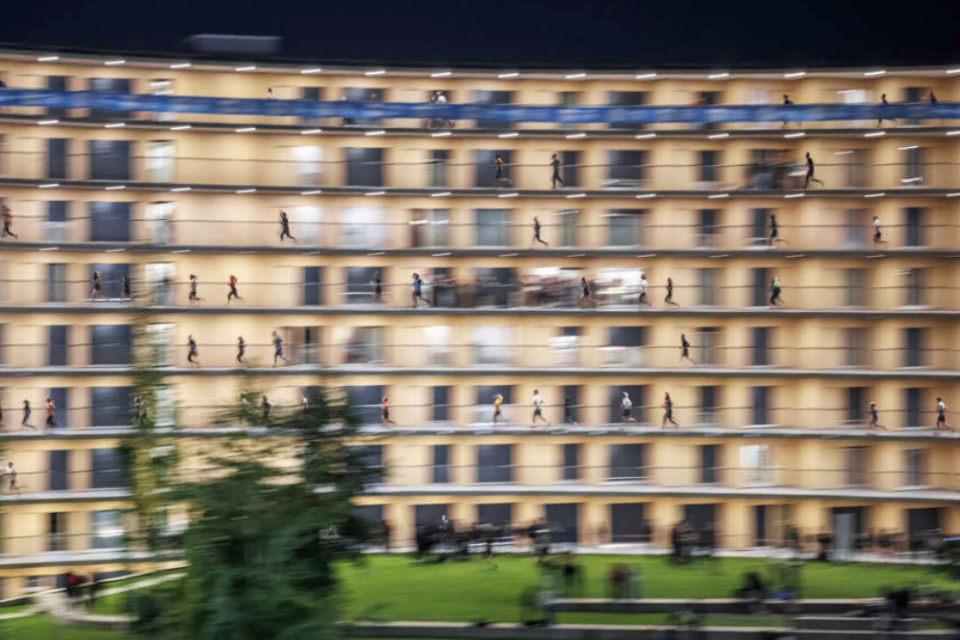 Ein unscharfes Bild eines mehrstöckigen Wohngebäudes bei Nacht, in dem zahlreiche Menschen entlang der Außenflure gehen. Im Vordergrund ist etwas Grün zu sehen.