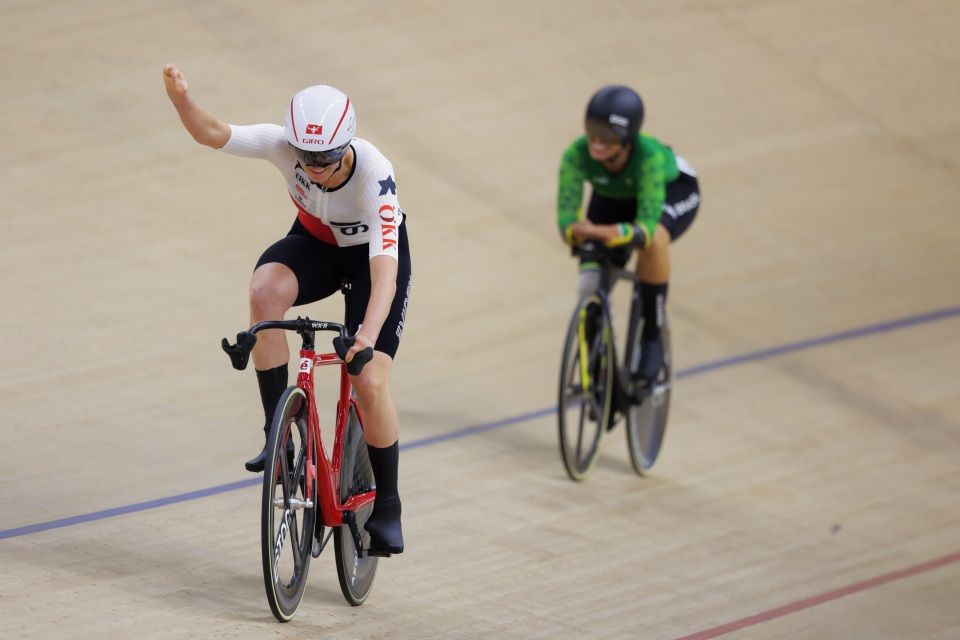 Ein Radfahrer in einem weiß-roten Trikot hebt jubelnd die Faust, während er auf einer Indoor-Bahn fährt. Ein anderer Radfahrer in einem grün-schwarzen Trikot folgt ihm.