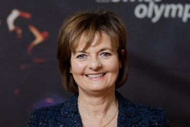 Eine Frau in einem dunklen Blazer lächelt vor einer Kulisse von Swiss Olympic, die eine Fackel und das Logo von Swiss Olympic zeigt.