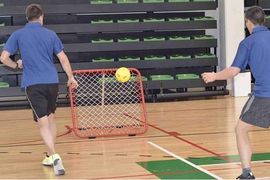 Zwei Personen in blauen Hemden spielen ein Hallenspiel, bei dem sie einen gelben Ball auf einen roten Abpraller mit Netz werfen. Sie befinden sich auf einem Sportplatz mit grünen und schwarzen Tribünen im Hintergrund.
