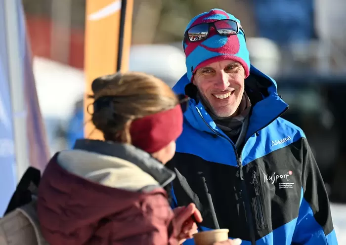 Ein Mann in blauer Winterjacke und bunter Mütze lächelt, während er an einem sonnigen Wintertag im Freien mit einer Frau mit kastanienbraunem Stirnband und Jacke spricht. Der Mann hält eine Tasse in der Hand und trägt eine Sonnenbrille auf seiner Mütze.